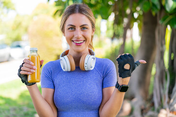 Wall Mural - Young pretty sport woman holding an orange juice at outdoors pointing to the side to present a product