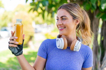 Wall Mural - Young pretty sport woman holding an orange juice at outdoors with happy expression