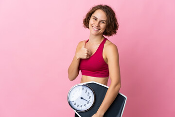 Young English woman isolated on pink background holding weighing machine with thumb up