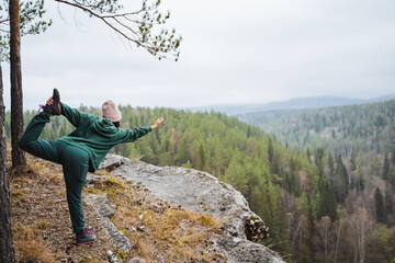 Woman doing yoga asana standing on the edge of a mountain with forest in the background, standing on one leg practice, meditation in nature, yoga travel, tour