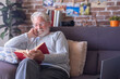© luciano - Smiling senior bearded man with eyeglasses relaxing on sofa at home reading a book. Retirement lifestyle concept
