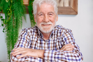 Wall Mural - Portrait of handsome smiling bearded senior man with crossed arms sitting at cafe table. Elderly caucasian male wearing checkered shirt looking at camera