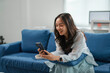 © amnaj - Young woman is sitting on a blue sofa at home, smiling while using her smartphone