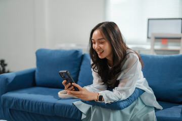 Young woman is sitting on a blue sofa at home, smiling while using her smartphone