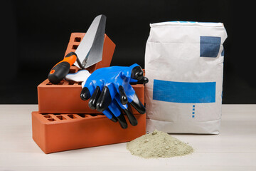 Poster - Red bricks, cement powder, putty knife and gloves on light wooden table against black background. Building materials