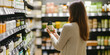 © laddawan - A woman in a cozy sweater examines a bottle of vitamins attentively while standing in a health supplement aisle, highlighting wellness and informed shopping choices.
