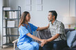 © amnaj - Young female nurse wearing blue scrubs is comforting a senior male patient on the sofa during a home visit