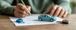 © Cheewynn - Close-up of man signing a car insurance contract with a blue toy car model on documents, car keys on table, vehicle purchase or warranty, professional setting, blurred background