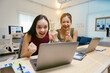© Parichat - Two businesswomen are sitting at their desks in the office, looking excitedly at a laptop screen and celebrating their success