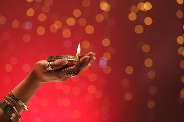 Sticker - Diwali celebration. Woman holding lit diya lamp on red background with blurred lights, closeup. Space for text