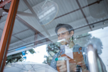 male waiter wearing apron cleaning glass window with wiper and spray at coffee shop