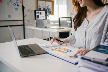 Poster - Businesswoman is analyzing financial data using a calculator and working on a laptop, with charts and graphs at her office desk