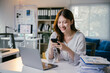 © PaeGAG - Young asian businesswoman smiling and using smartphone while working on laptop computer with paperwork on desk in modern office, concept of technology and communication