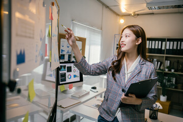 Poster - Young businesswoman is holding a clipboard and pointing at charts on a whiteboard during a presentation in the office