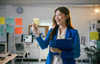 © PaeGAG - Young asian businesswoman happily smiles while holding a sticky note in a vibrant office, embodying a modern and professional approach to work