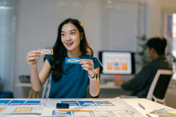 Poster - Young asian woman working as ui/ux designer is smiling while holding two colorful paper prototypes during a brainstorming session