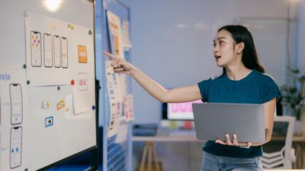 Poster - Young professional web designer is holding a laptop and pointing at a whiteboard covered with wireframes, explaining her work