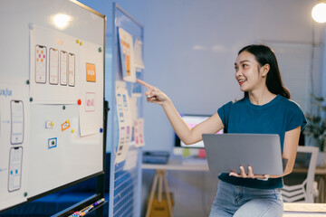 Poster - Young professional woman is standing in her office, holding a laptop and pointing at a whiteboard with mobile app design sketches on it