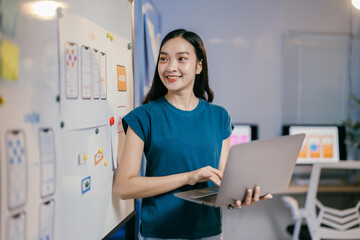 Poster - Young asian designer woman happily holds a laptop while working on a new mobile app in her office, fully engaged in the creative process