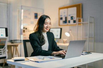 Poster - Young asian businesswoman is working late in her office and smiling at her laptop. She is wearing a black blazer and a white shirt