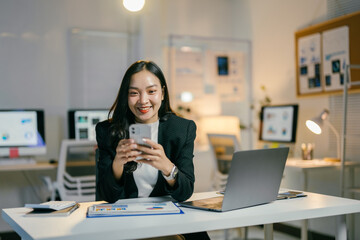 Young businesswoman is using her smartphone while sitting at her desk in a modern office. She is smiling and appears to be enjoying her work