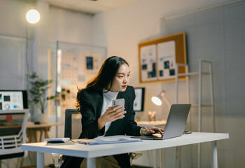 Poster - Young professional sitting at her desk in a modern office, working late and using her laptop and smartphone at the same time