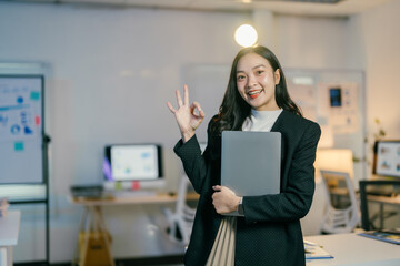 Confident asian businesswoman smiling in modern office, holding laptop and making ok sign gesture, exuding success and happiness in work environment