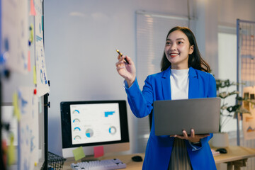 Fototapete - Young professional woman is standing in her office holding a laptop and pointing at graphs and charts on a whiteboard during a business presentation