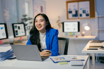 Poster - Businesswoman wearing a blue blazer sits at her desk working on a laptop with a smile on her face, surrounded by paperwork and digital screens