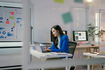 Poster - Young asian businesswoman is working on her laptop in a modern office, surrounded by charts and graphs. She is concentrating on her work and is determined to succeed