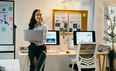 Young professional woman holding a laptop while leaning on a desk in a modern office, surrounded by computers and whiteboards with data visualizations