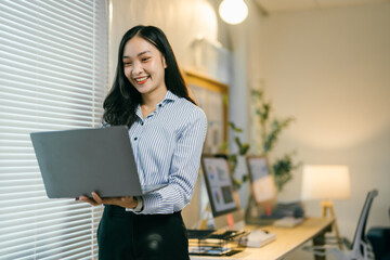 Asian businesswoman happily works from her office, confidently holding her laptop, showcasing her professional and focused demeanor in a modern setting