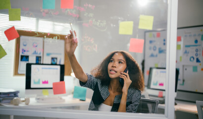 Aufkleber - Young manager multitasks in a modern office, brainstorming project ideas while writing on a glass wall and talking on the phone, exuding determination and confidence in her corporate setting