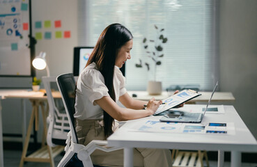 Poster - Asian businesswoman is analyzing financial graphs on a clipboard while sitting at her desk in the office. She is working on a new project