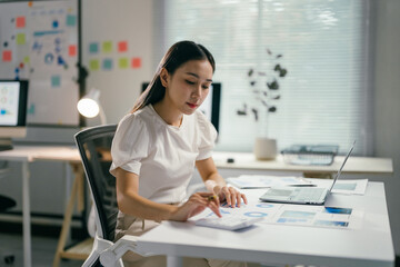 Poster - Focused young asian businesswoman analyzing financial data with a calculator in a modern office, surrounded by paperwork, charts, and graphs, using technology to excel in her career