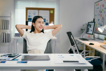 Aufkleber - Young asian businesswoman is leaning back in her chair with her hands behind her head, taking a break from working on a laptop at her desk in a modern office