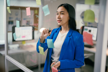 Poster - Young manager is using colorful sticky notes and a marker to brainstorm ideas on a transparent glass wall in her office