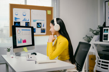 Aufkleber - Young professional is working in her office at her desk. She is wearing headphones and smiling at her desktop computer screen