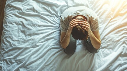 young man is sitting on his bed, holding his head in his hands, suffering from a headache. the brigh