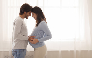 Happy young couple expecting baby standing together against window at home, loving husband tenderly touching belly of his pregnant wife, free space