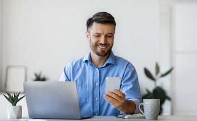 Poster - Handsome male office manager using smartphone at workplace, sitting at desk, checking social media or browsing app, answering to message, having break, looking at screen and smiling, copy space
