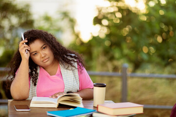Poster - A student sits at a table surrounded by books, looking thoughtfully while holding a pen. She is outdoors in a sunny environment, deep in contemplation about her studies.