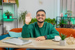 © Andrii Iemelianenko - Hello. Indian smiling man smiling friendly at camera and waving hands gesturing hello hi, greeting or goodbye welcoming with hospitable expression at home office. Arabian guy sitting at table in room