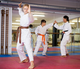 Wall Mural - Sporty girl in kimono posing in gym while other kids sparring in background during karate class