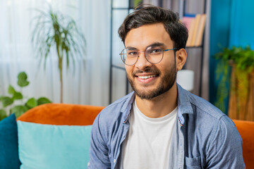 Portrait of happy calm Indian Arab man at home couch smiling friendly glad expression looking away dreaming resting relaxation feel satisfied good news celebrate win. Arabian young guy in living room