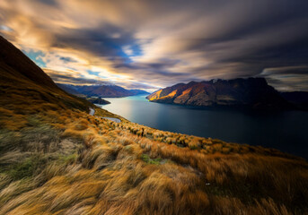 Poster - A winding road leads down to a serene lake surrounded by snow capped mountains under a sky with dramatic clouds.