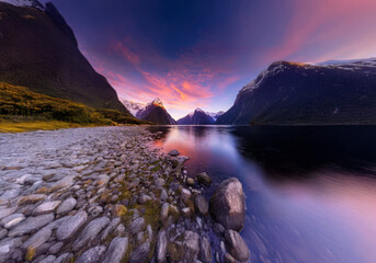 Poster - A scenic view of a lake with mountains in the background at sunset. The sky is a vibrant pink and the water is a calm blue.