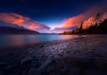 Aufkleber - A serene landscape features a tranquil lake, a shoreline of smooth stones, and snow-capped mountains against a vivid sunset sky.