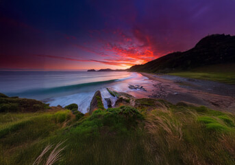 A vibrant sunset paints the sky with streaks of red and pink over a coastline with a gentle wave breaking on the shore.