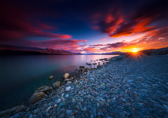 Poster - Pink and orange clouds fill the sky above a calm lake and distant snow-capped mountains.